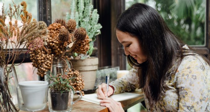 happy young ethnic woman writing article in cozy workplace